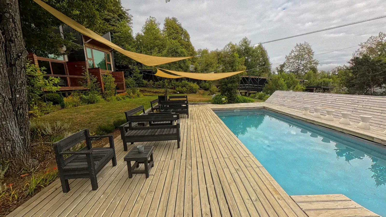 Piscina de Ni-Newen Lodge con terraza de madera, sillones de exterior y uno de los edificios del hotel entre el bosque, en su recorrido virtual 360